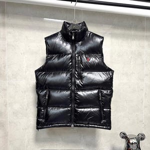 Puede incluir: Un chaleco acolchado negro brillante con cuello alto y cremallera completa. El chaleco tiene dos bolsillos laterales y un pequeño logotipo rojo en el pecho. Cuelga de una percha metálica.