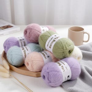 May include: A selection of pastel-coloured knitting yarn balls, including lavender, pink, green, and blue. Each ball is labelled with "Snowflake Yarn Knitting Yarn". The yarn is arranged on a wooden tray, with a mug and a grey blanket in the background.