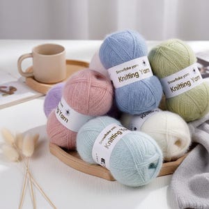Puede incluir: Una colección de ovillos de lana para tejer en tonos pastel, incluyendo azul, rosa, verde y blanco. Cada ovillo tiene una etiqueta blanca que dice "Snowflake Yarn Knitting Yarn". La lana está dispuesta sobre una bandeja de madera.