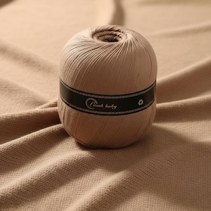 Puede incluir: Una madeja de hilo color beige con una etiqueta negra que dice "Lamb baby". El hilo está sobre una tela de punto beige. El hilo está bien enrollado y tiene un agujero en el centro.