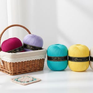 May include: A wicker basket filled with purple and fuchsia yarn balls, with a teal, yellow, and white yarn balls next to it. Each ball has a black band with white text. A crocheted square is in the foreground.