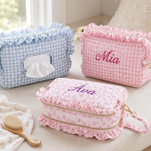 May include: Three rectangular fabric pouches with ruffled edges and gold zips. One blue gingham pouch features a central wipe dispenser. A pink gingham pouch is embroidered with the name Mia. A floral print pouch is embroidered with the name Ava.