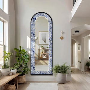 Puede incluir: Un espejo de pie arqueado con un marco decorativo adornado con un patrón de azulejos azules y blancos. El espejo tiene un borde exterior negro fino y una superficie de vidrio reflectante que muestra un interior con estantería y arte.