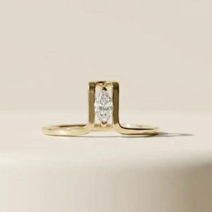 Peut inclure: Une bague en or avec un diamant central de taille marquise. La bague présente un design unique avec une barre verticale sertissant le diamant et un anneau incurvé. La bague est posée sur un fond neutre.