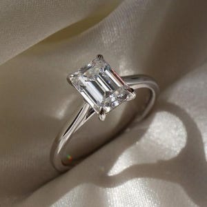 Pode incluir: Um anel de prata com uma grande pedra preciosa clara de forma retangular. O anel está sobre um tecido branco e sedoso, com a luz criando uma sombra do anel. A pedra tem um design de corte em degraus.