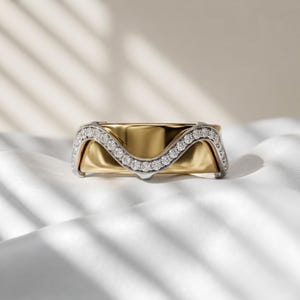 Könnte beinhalten: Ein zweifarbiger Ring mit einer glatten, polierten Gelbgoldschiene und einer wellenförmigen silberfarbenen Auflage. Der silberfarbene Teil ist mit einer Reihe kleiner, klarer, rund geschliffener Steine besetzt, die dem geschwungenen Verlauf folgen.