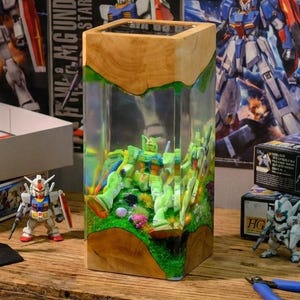 May include: A wooden and clear resin display case featuring miniature Gundam figures. The case sits on a wooden table with other Gundam model kits and tools. The background includes Gundam posters and boxes.