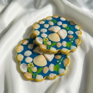 Puede incluir: Dos posavasos de resina con fondo azul, incrustados con conchas marinas y pequeñas piedras. Los posavasos tienen un borde festoneado con un ribete dorado. Hojas verdes y flores amarillas están pintadas en la superficie.