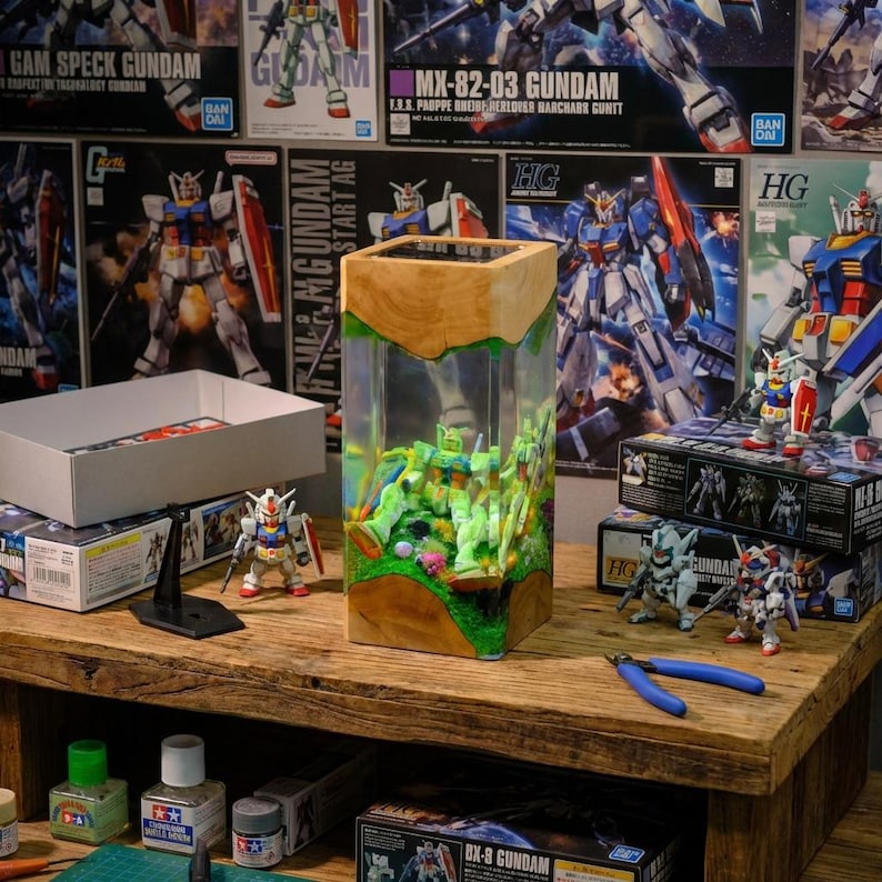 May include: A wooden and clear resin display case featuring miniature Gundam figures. The case sits on a wooden table with other Gundam model kits and tools. The background includes Gundam posters and boxes.