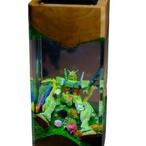 May include: A wooden block with a clear resin interior encases a green and yellow robot figure. The robot is positioned on a bed of green and pink miniature plants. The wooden exterior has a natural, light brown color.