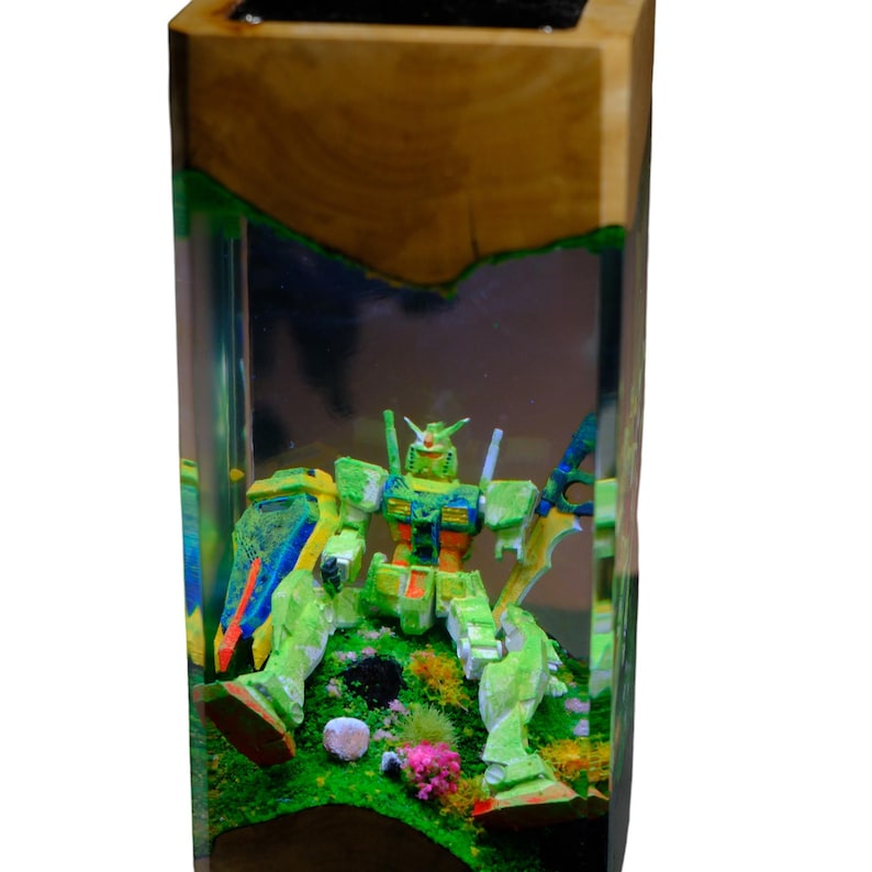 May include: A wooden block with a clear resin interior encases a green and yellow robot figure. The robot is positioned on a bed of green and pink miniature plants. The wooden exterior has a natural, light brown color.