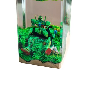 May include: A clear resin block encasing a green and blue robot figure amidst vibrant green foliage. The robot has red accents on its feet. The base of the block is a light-colored wood.