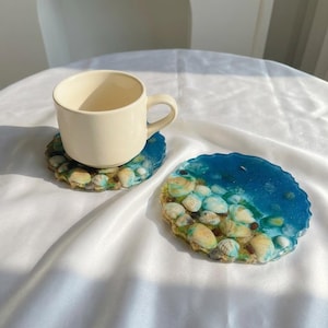 Puede incluir: Dos posavasos de resina con temática de playa. Uno de los posavasos sostiene una taza de color crema. Los posavasos presentan un diseño de resina azul y transparente con conchas marinas y arena incrustadas, creando una estética costera. Los posavasos son redondos.