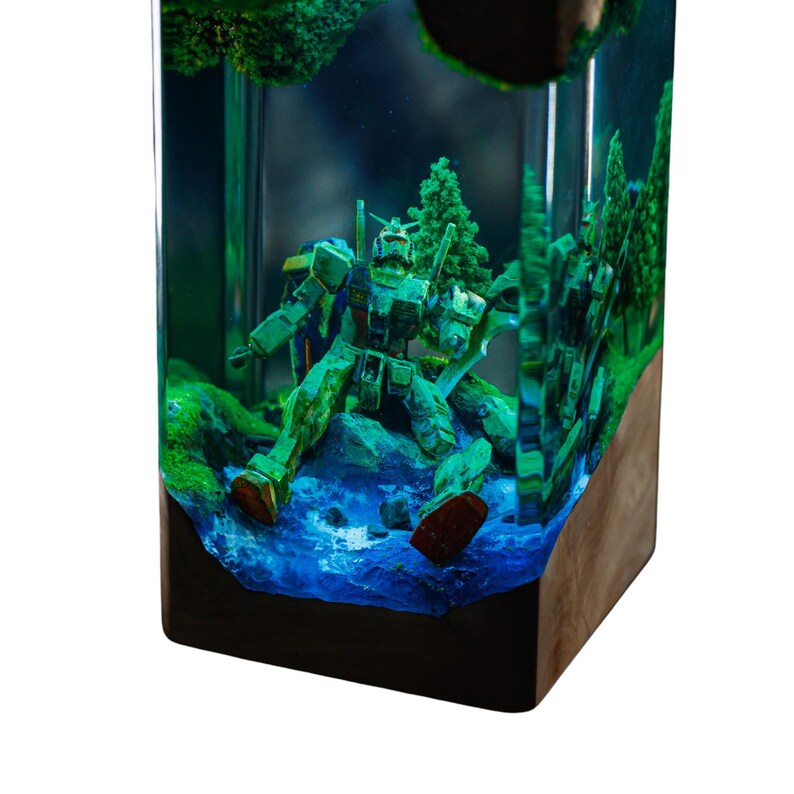 May include: A green and brown robot figure is encased in a clear resin block, set against a miniature landscape with blue water and green foliage. The block has a dark wooden base and a dark top.