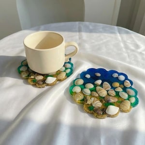 Puede incluir: Dos posavasos de resina con conchas incrustadas y una taza crema. Los posavasos presentan un diseño azul, verde y amarillo. Un posavasos tiene una taza encima. Los posavasos miden aproximadamente 10 cm de diámetro.