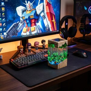 May include: A desktop setup featuring a computer monitor displaying a Gundam image, a mechanical keyboard, and small anime figurines. A unique resin art piece with a miniature landscape is on the desk, alongside a mouse and headphones.