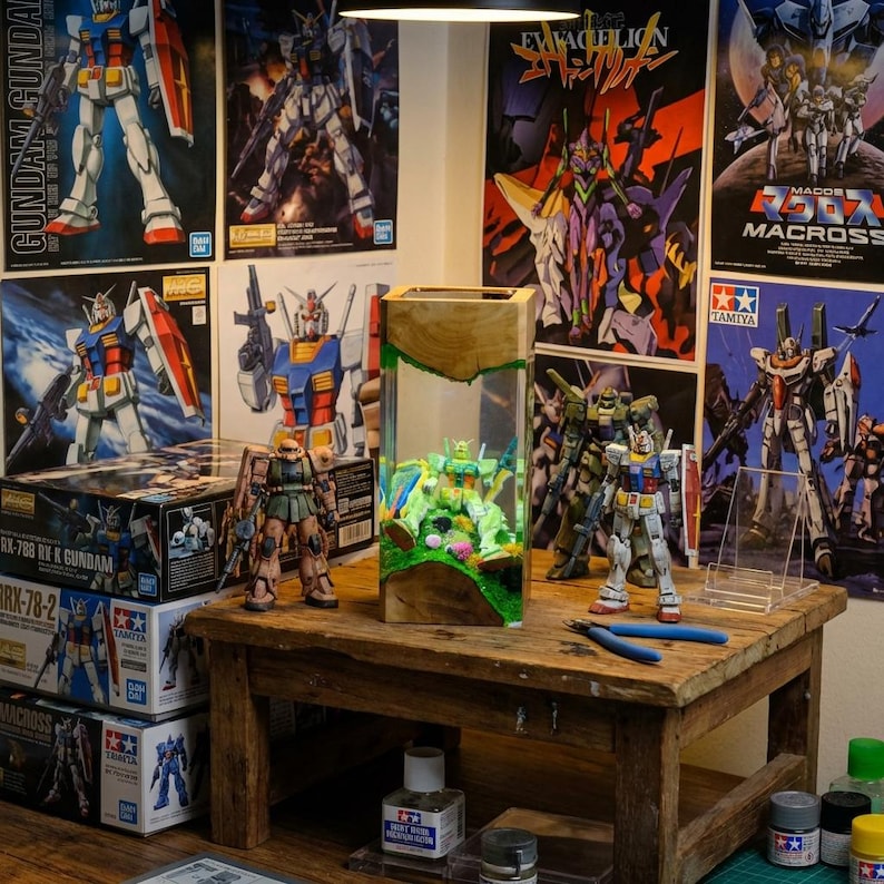 May include: A wooden table displays several Gundam model kits and assembled figures. A clear display case houses a miniature diorama. Posters of anime characters and model kit boxes are visible in the background. Tools and paint bottles are also on the table.