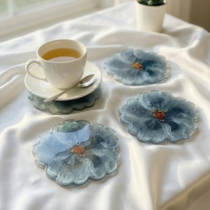 Puede incluir: Una taza y un platillo blancos con una cuchara plateada descansan sobre una pila de cuatro posavasos con forma de flor. Los posavasos son azules con un centro dorado. Una pequeña planta suculenta en una maceta blanca está en el fondo.