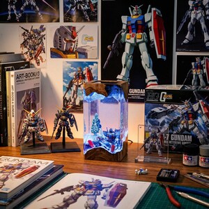 May include: A wooden-framed, rectangular lamp with a miniature diorama inside, featuring a Gundam model, trees, and a blue-lit background. The scene is surrounded by Gundam model kits, posters, and art books, creating a collector's display.