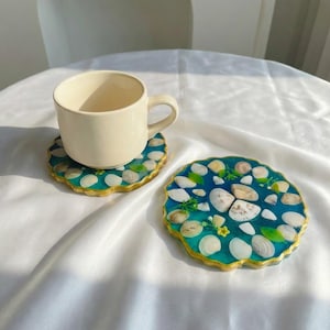 Puede incluir: Dos posavasos de resina con diseño de conchas marinas. Los posavasos tienen un fondo azul con conchas incrustadas, detalles verdes y un borde dorado. Una taza crema está encima de uno de ellos.
