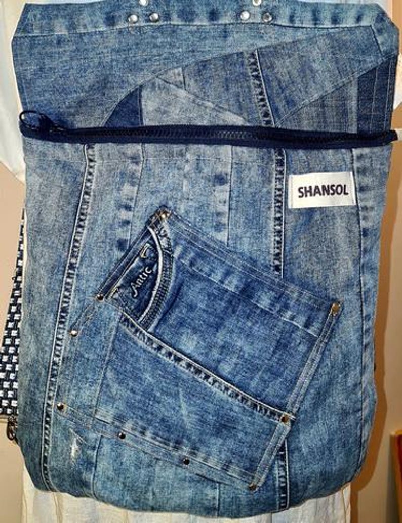 May include: A blue denim backpack with a front pocket and a zipper closure. The backpack is made from recycled denim and features the brand name "SHANSOL" on a white label. The front pocket has the word "Antic" on it.