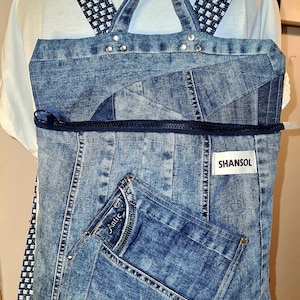 May include: A denim backpack with a rectangular shape and a front pocket made from a pair of jeans. The backpack has a zipper closure and a label that reads "SHANSOL". The straps are patterned with a white and navy blue design.