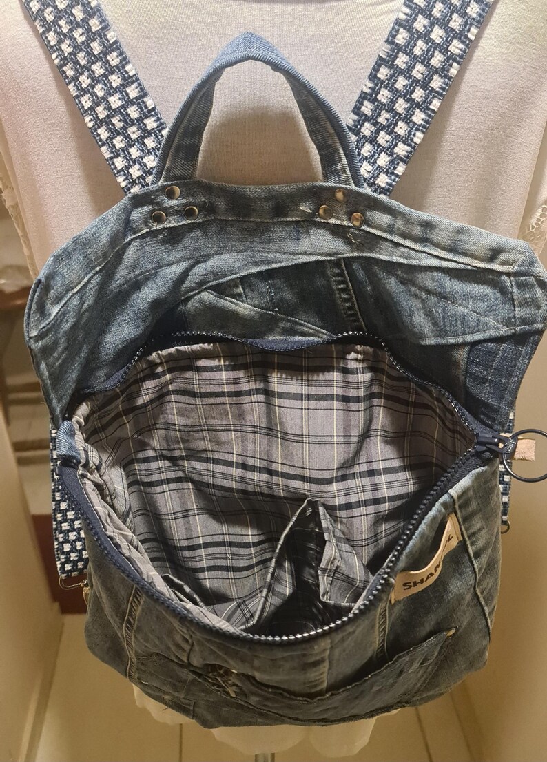 May include: A denim backpack with a plaid interior and a zipper closure. The straps are a blue and white checkered pattern. The bag has a top handle and a small pocket on the front. The interior is gray and black plaid.