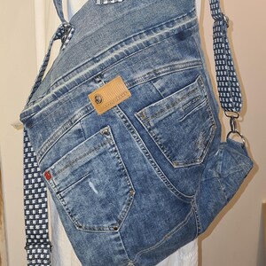 May include: A denim backpack made from recycled jeans. The bag features the back pockets of the jeans, a brown leather tag, and adjustable straps with a black and white checkered pattern. The bag is a unique, eco-friendly accessory.