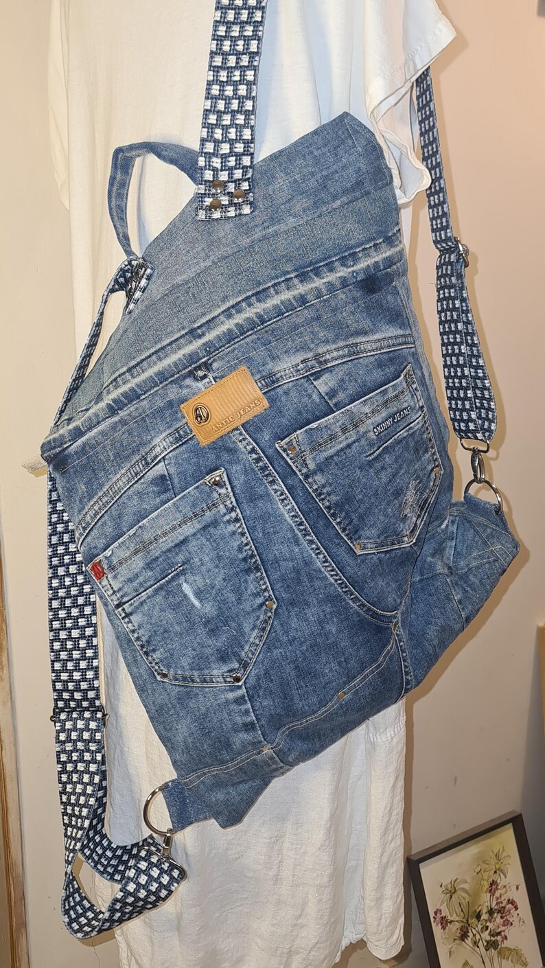 May include: A denim backpack made from recycled jeans. The bag features the back pockets of the jeans, a brown leather tag, and adjustable straps with a black and white checkered pattern. The bag is a unique, eco-friendly accessory.