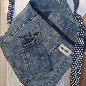 May include: A denim backpack with a zipper closure and a small front pocket. The backpack has a blue and white checkered strap and the brand name "SHANSOL" is visible on a tag. The word "Antic" is embroidered on the pocket.