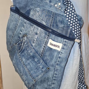May include: A denim backpack with a blue and white patterned strap. The bag is made from light blue denim with a zipper closure and a small pocket. The brand name "SHANSOL" is visible on a white tag.