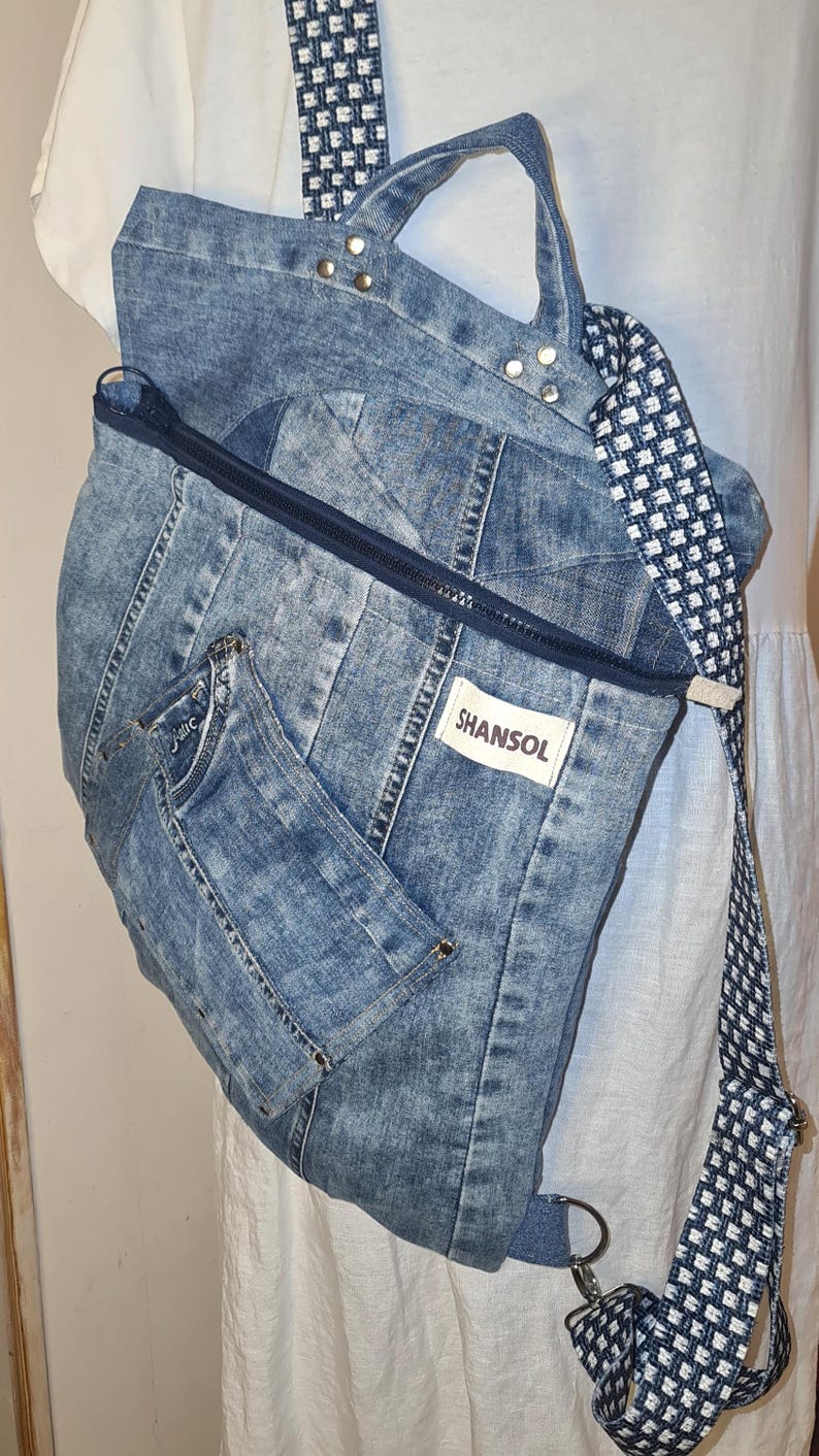 May include: A denim backpack with a blue and white patterned strap. The bag is made from light blue denim with a zipper closure and a small pocket. The brand name "SHANSOL" is visible on a white tag.