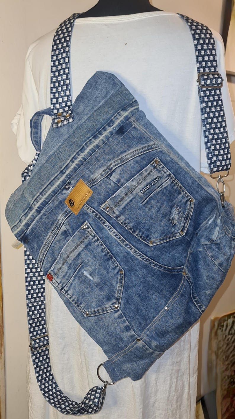 May include: A denim backpack made from recycled jeans. The bag features the pockets and seams of the jeans, with a brown leather tag and a patterned strap. The bag is a unique, eco-friendly accessory.