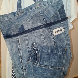 May include: A denim backpack with a blue and white checkered strap. The backpack is made from recycled jeans and features a front pocket, a zipper closure, and the brand name "SHANSOL". The bag has silver rivets and a small keychain.