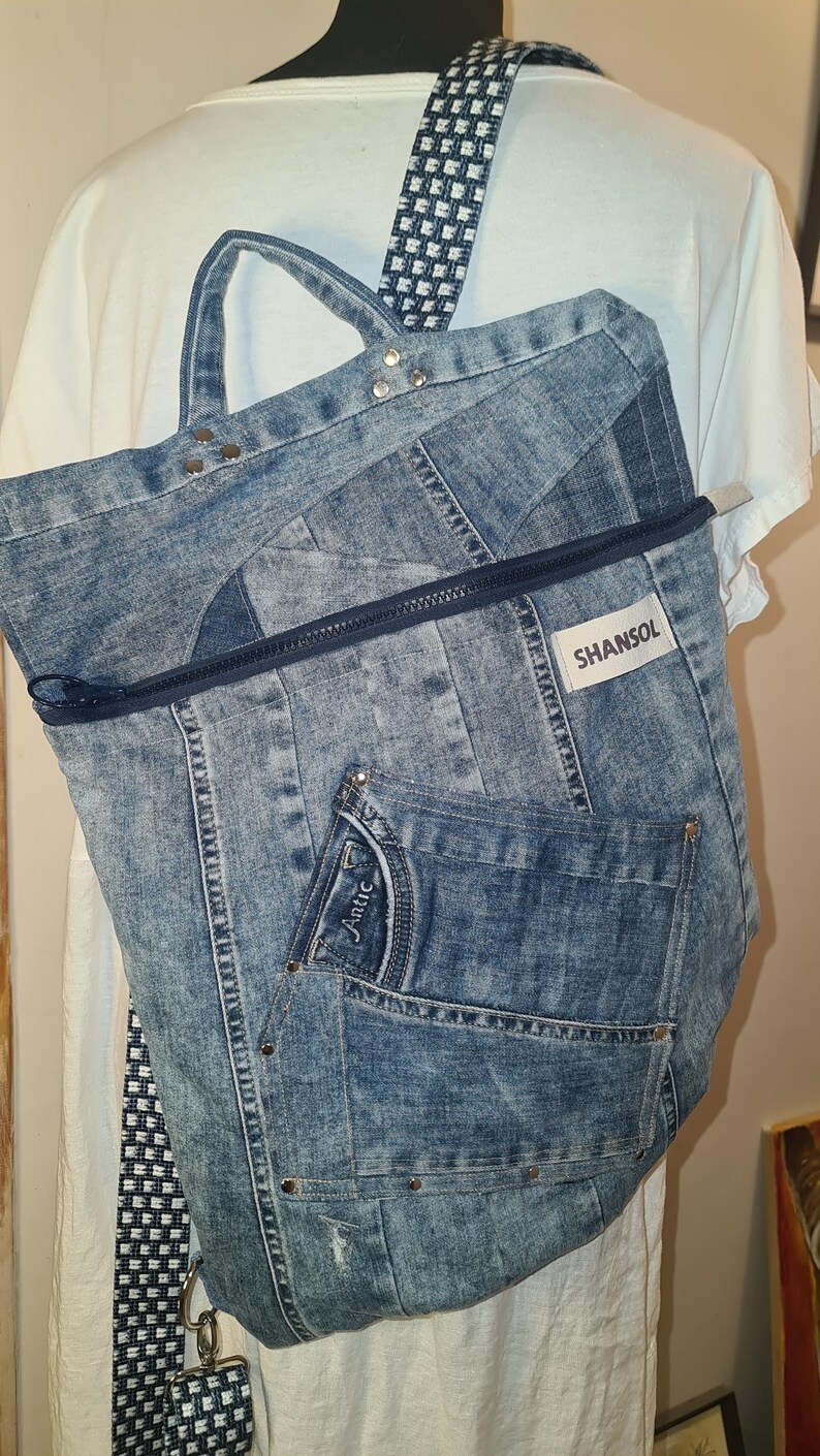 May include: A denim backpack with a blue and white checkered strap. The backpack is made from recycled jeans and features a front pocket, a zipper closure, and the brand name "SHANSOL". The bag has silver rivets and a small keychain.