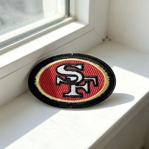 Może przedstawiać: Owalna, haftowana naszywka z logo San Francisco 49ers. Wzór zawiera biały, spleciony monogram SF na czerwonym tle, otoczony cienką złotą obwódką oraz grubym, czarnym, obszytym brzegiem zewnętrznym.