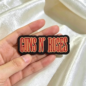 May include: A black and red embroidered patch with the text "GUNS N' ROSES" in a stylized font. The patch has a black border and is held against a white, textured background.