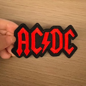May include: A fabric patch featuring the text AC/DC in bright red embroidery with a thick black border. The letters are stylized with sharp, angular edges, and a red lightning bolt symbol separates the letters AC and DC in the center.