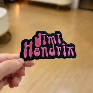 May include: An embroidered patch featuring the text Jimi Hendrix in bright pink thread against a solid black background. The lettering has a stylized, rounded font, and the entire patch is outlined in a thick black border with a scalloped edge.