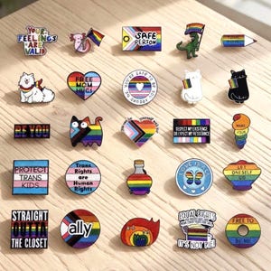 以下が含まれることがあります： 虹、猫、"Safe Person"や"Trans Rights are Human Rights"などのフレーズを含む、さまざまなデザインとテキストが施されたカラフルなエナメルピンのコレクション。 ピンは明るい木製の表面に配置されています。