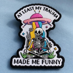 Può includere: Toppa ricamata con uno scheletro, un'astronave e la scritta "AT LEAST MY TRAUMA MADE ME FUNNY". L'astronave emette un raggio arcobaleno sullo scheletro. Bordo nero.