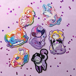 Puede incluir: Una colección de siete parches bordados de gatos en varios diseños. Los parches presentan gatos como sirenas, brujas y otros personajes caprichosos. La paleta de colores incluye rosas, morados, naranjas y azules. Los parches están esparcidos sobre una superficie morada clara con confeti en forma de corazón.