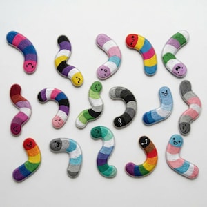May include: A collection of colorful, embroidered worm-shaped patches. Each patch features a unique combination of colors, including rainbow, pink, blue, green, and purple. The patches have small, embroidered faces and are arranged on a white surface.
