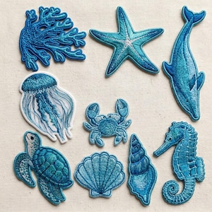 Op de afbeelding: Een verzameling geborduurde patches met verschillende zeedieren. Inclusief koraal, een zeester, een dolfijn, een kwal, een krab, een zeeschildpad, een zeeschelp, een conch en een zeepaardje. De patches zijn in blauw- en wittinten, met gedetailleerde steken.
