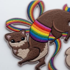 May include: Embroidered patch featuring a cartoon otter with a rainbow-colored sash and tail. The otter is brown with tan accents and has a playful expression. The rainbow colors include red, orange, yellow, green, blue, and purple.