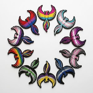 May include: A selection of embroidered phoenix patches in a variety of colours, including rainbow, pink, blue, and purple. Each patch has a black outline and represents different pride flags.