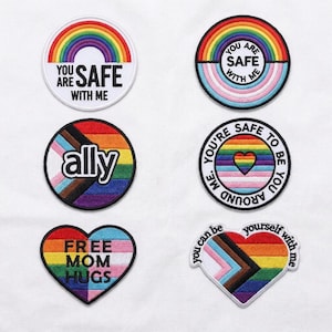 Könnte beinhalten: Eine Sammlung von gestickten Aufnähern mit Regenbogen- und LGBTQ+-Pride-Flaggen-Designs. Die Aufnäher zeigen Sätze wie "You Are Safe With Me", "Ally" und "Free Mom Hugs". Die Aufnäher sind rund und herzförmig.