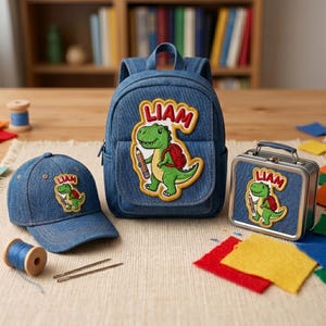 Peut inclure: Ensemble comprenant un sac à dos, une casquette et une boîte à lunch en jean. Chaque article présente un écusson avec un dinosaure vert tenant un crayon, un sac à dos rouge et le nom "LIAM" dans une bulle jaune. Pour enfants.