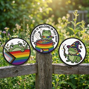 May include: Three round patches featuring cartoon frogs. One frog peeks over a rainbow flag, another stirs a pot of soup with the text "Everyone is welcome in this soup", and the last wears a wizard hat and cape.