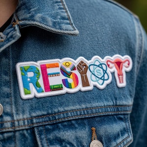 Könnte beinhalten: Eine blaue Jeansjacke mit einem weißen, bestickten Aufnäher mit der Aufschrift "RESIST". Der Aufnäher zeigt einen Globus, Regenbogenstreifen, eine geballte Faust, ein Wissenschaftssymbol und eine Gebärmutter.